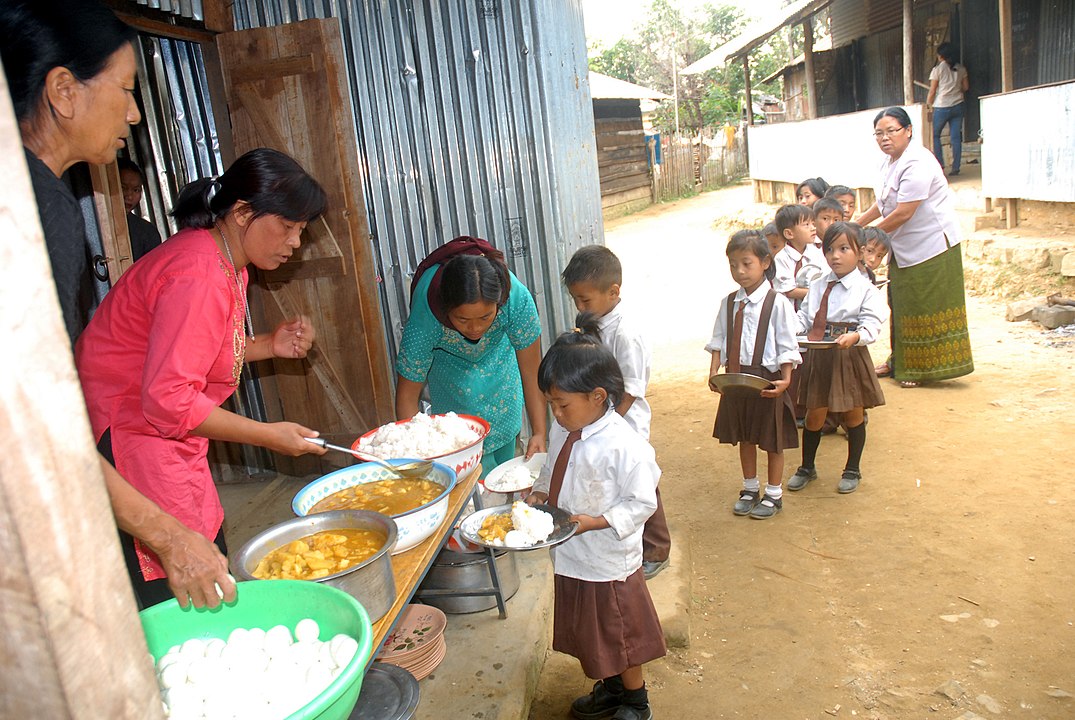 1075px-The_Children_being_served_the_food_under_the_Mid-day_Meal_Scheme_at_a_primary_school,_Wokha_district_in_Nagaland.jpg